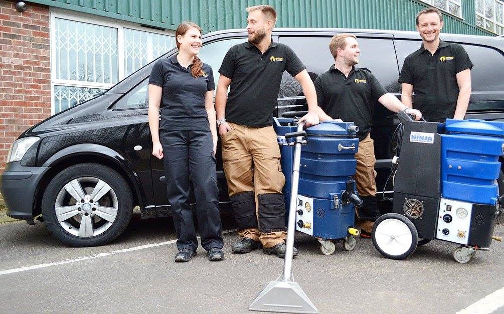 This is a photo of the team at The K&S Carpet  Cleaners. There are three men and one woman standing in front of their black van also with two steam carpet cleaners in front of them.